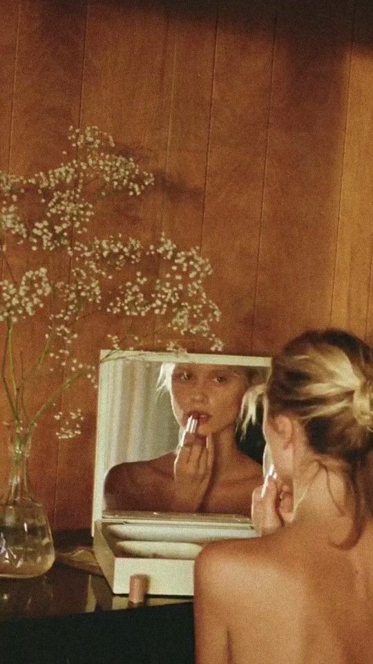 A woman applying lipstick in a small vanity mirror beside dried flowers
