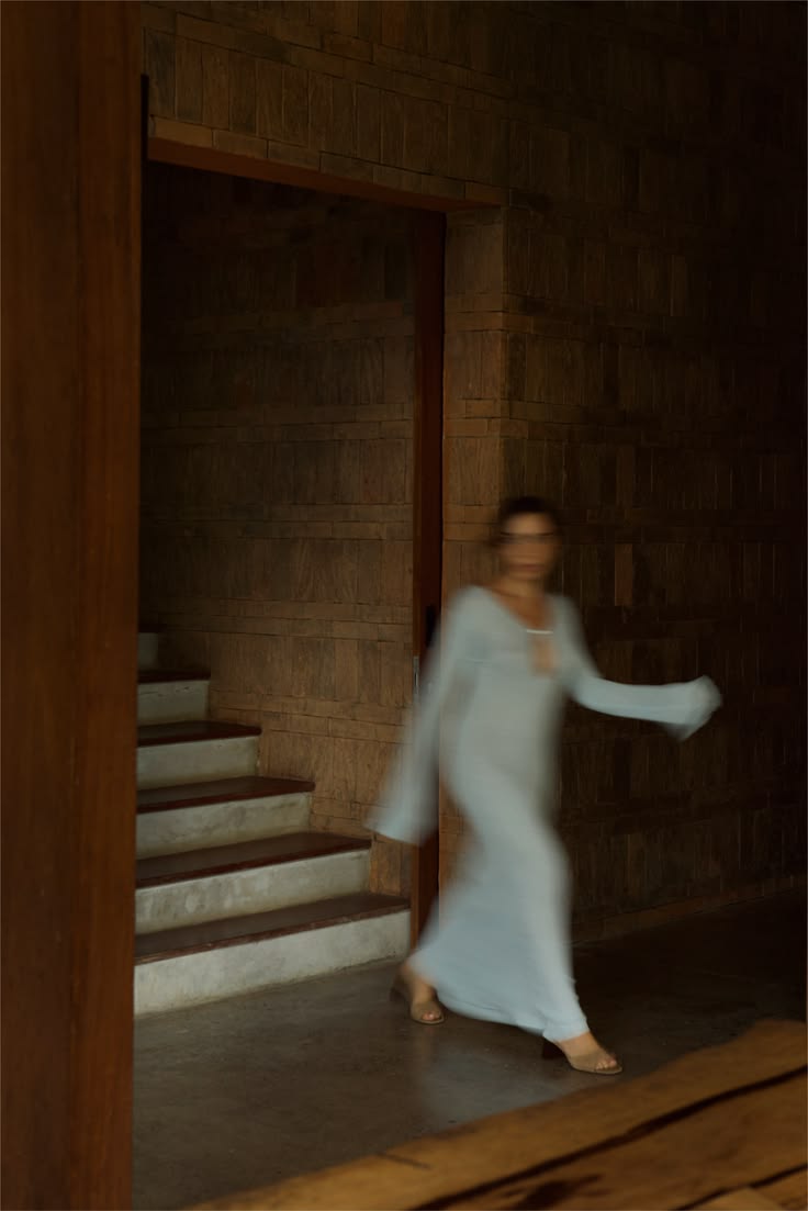 A blurred woman in a pale dress walking through a dim wooden hallway